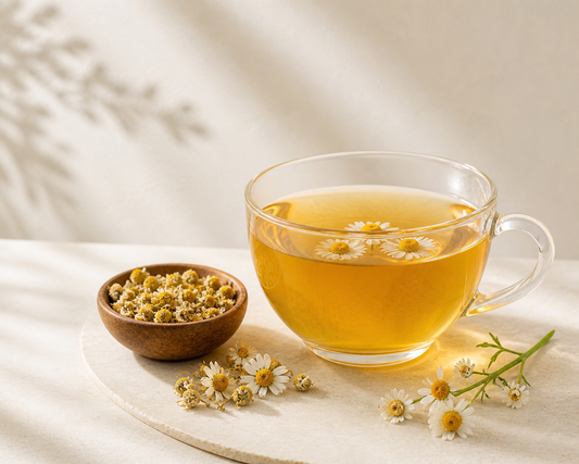 Clear glass mug filled with chamomile tea on a light surface with dried chamomile flowers and fresh chamomile blossoms.